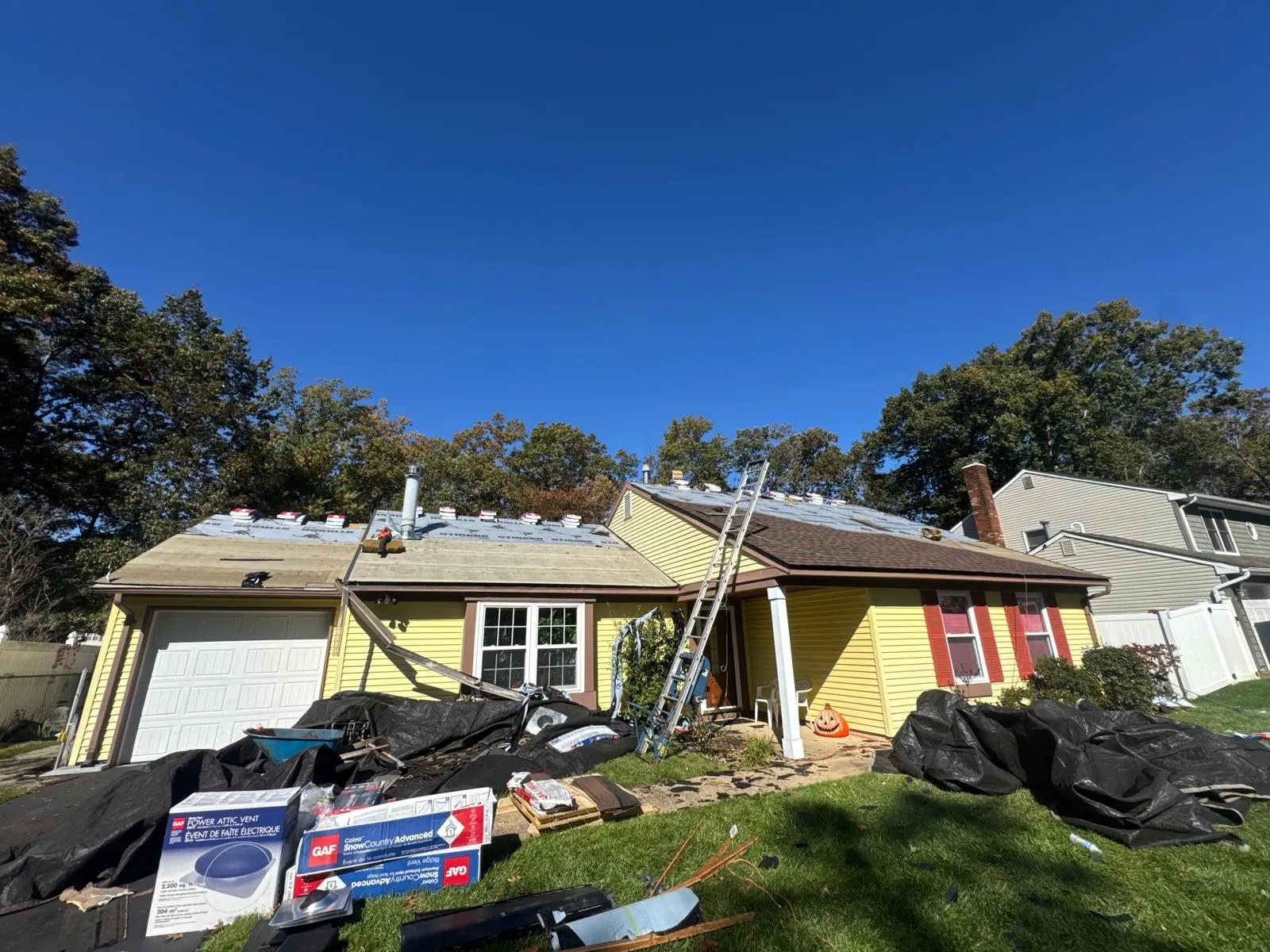 Roof tear-off in progress on a yellow house, with a ladder, tarps and GAF materials on the lawn